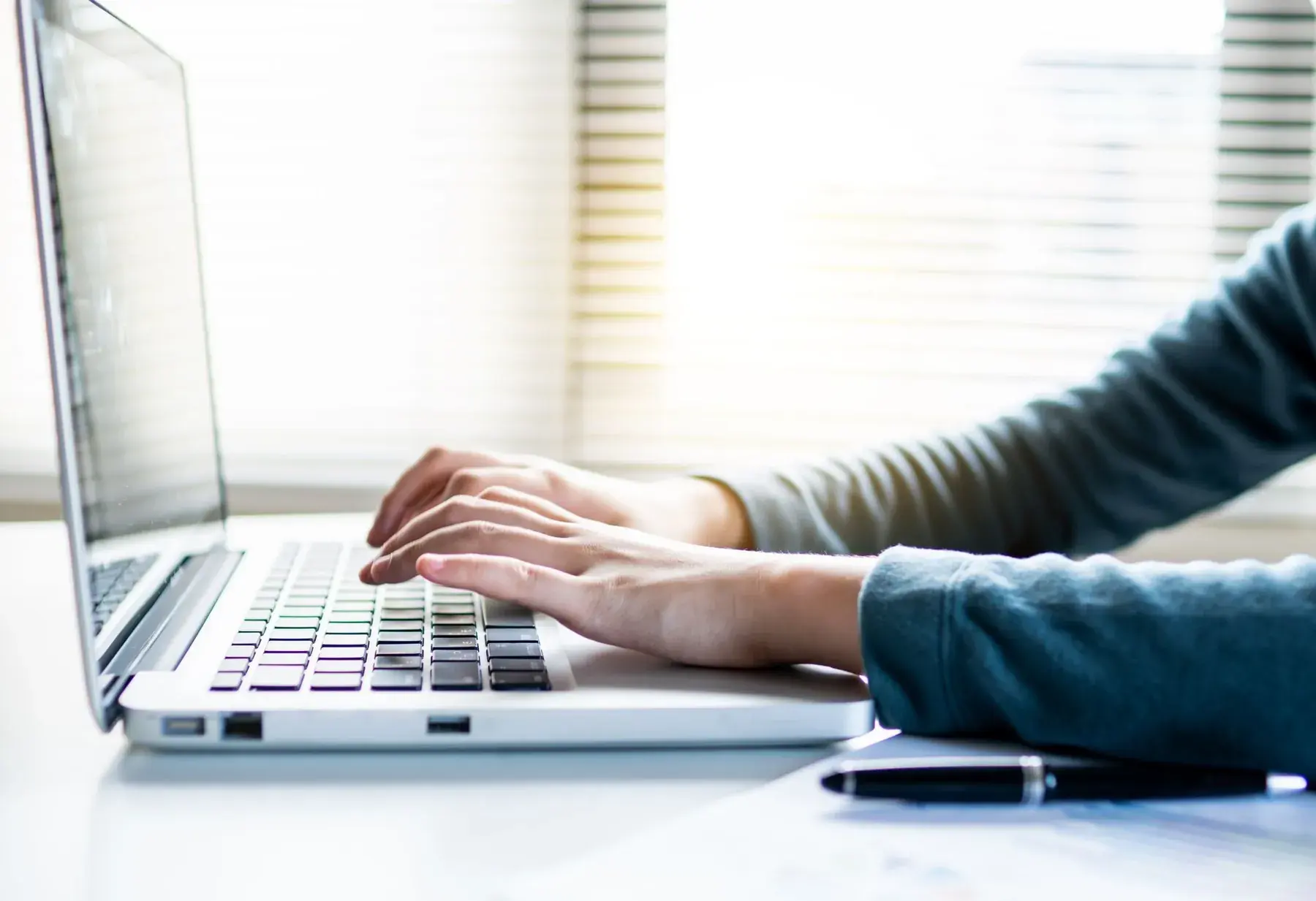Hands typing on a laptop by a window.