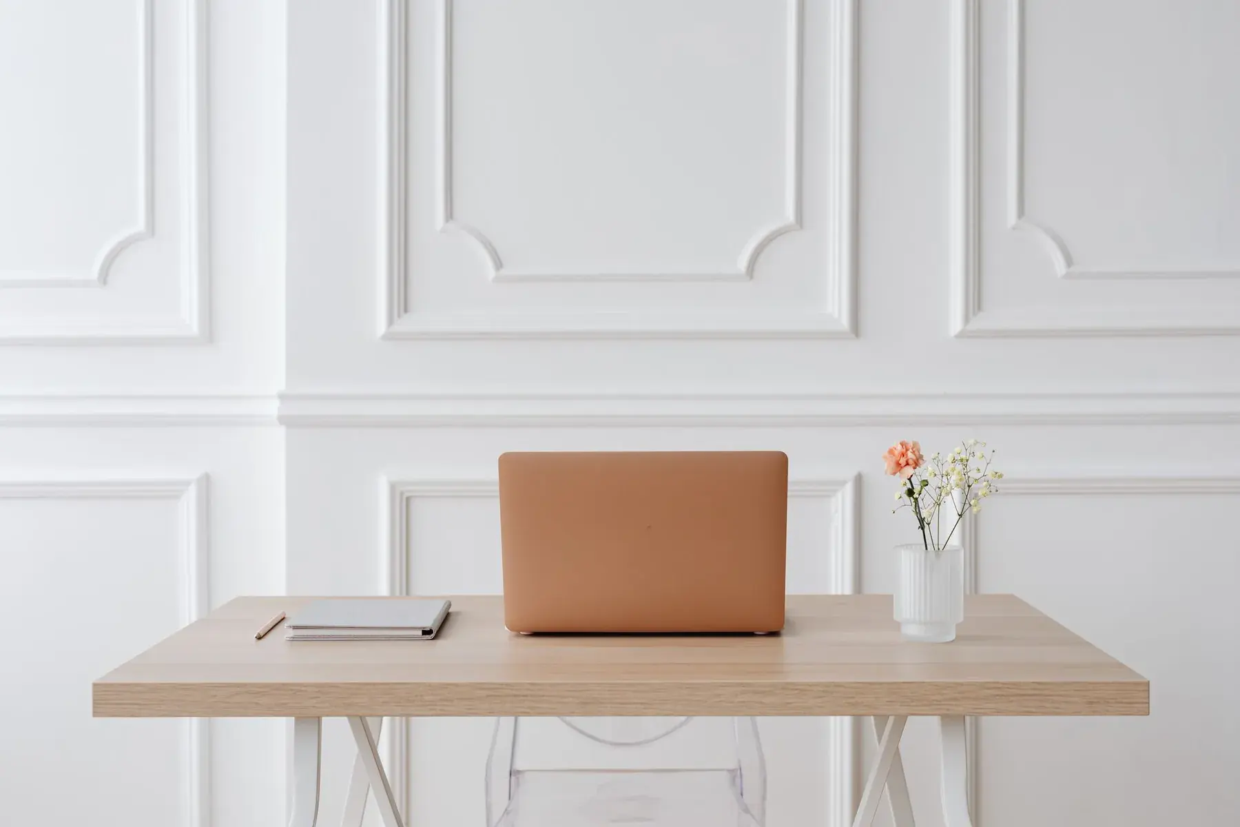 A simple desk with a laptop and flowers against a white wall.